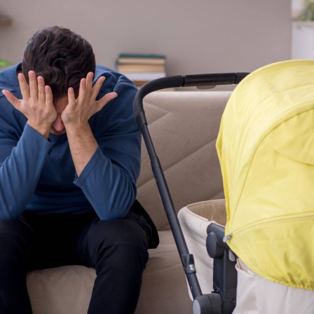 Ein junger Vater sitzt vor einem Kinderwagen und schlägt sich die Hände vor das Gesicht