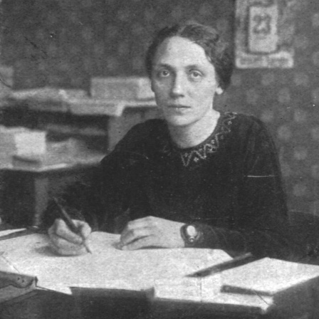 Historisches Schwarzweißfoto einer jungen Frau, die mit ernstem Ausdruck an ihrem Schreibtisch sitzt.
