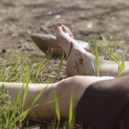 Am Wegesrand liegt das Opfer eines Serienmörders. Perspektive auf einen Rockansatz und nackte Beine. Am linken Fuß trägt das Opfer einen Halbschuh, der rechte Fuß ist blutbeschmiert und nackt.