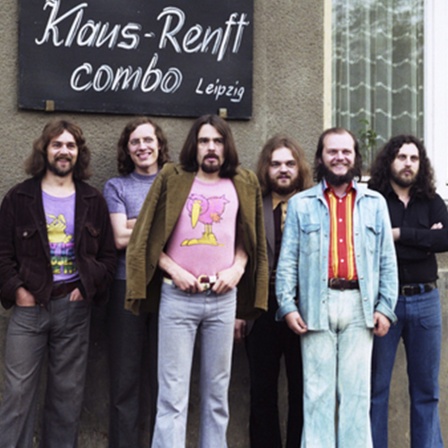 Die Klaus-Renft-Combo posiert vor einer grauen Hauswand, auf dem ein Schild angebracht ist mit der Aufschrift "Klaus Renft Combo Leipzig".