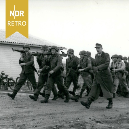 Mit einem Lied marschiert eine Gruppe des Bundesgrenzschutzes vorbei an einer der ersten fertigen Hütten, 1954