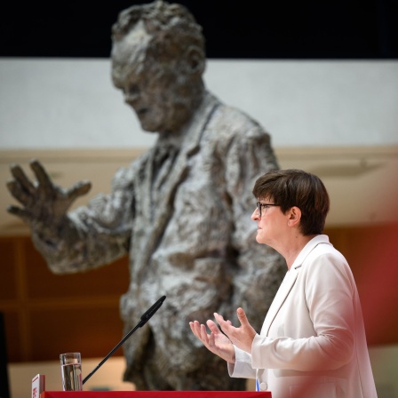 Saskia Esken gestikuliert am Rednerpult vor einer überlebensgroßen Skulptur von Willy Brandt. 