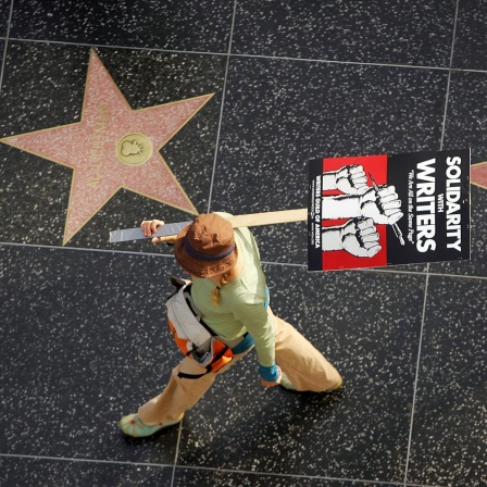 Aufnahme aus der Vogelperspektive zeigt eine Gewerkschaftsmitglied mit einem Plakat auf dem steht "Solidarity with Writers" zwischen zwei Sternen auf  dem Hollywood Walk of Fame.