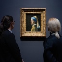 Zwei Frauen stehen vor einem Gemälde und betrachten es. Das Bild hängt an einer dunklen Wand. Es ist das Gemälde "Mädchen mit dem Perlenohrgehänge" des niederländischen Malers Jan Vermeer. Es zeigt ein junge Frau im Halbprofil mit einem hell leuchtenden Perlenohrring.