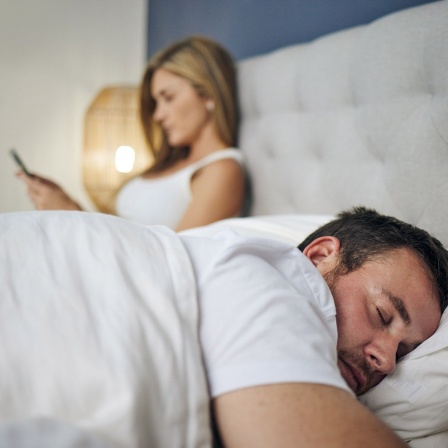 Ein Mann schläft im Doppelbett - seine Frau sitzt neben ihm und hat das Handy in der Hand. 
