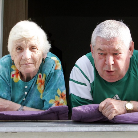 Ein älteres Ehepaar schaut, auf Kissen gelehnt,  aus dem Fenster