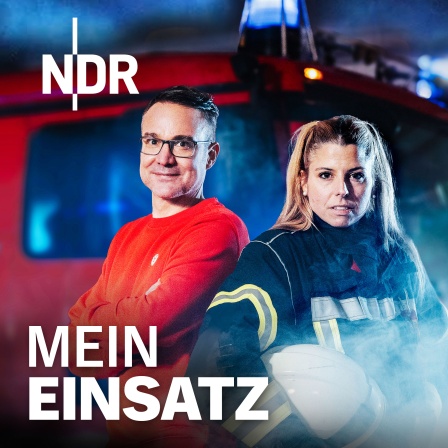 Ein Mann in rotem Pullover und eine Frau in Feuerwehruniform mit Helm stehen vor einem roten Feuerwehrauto mit Blaulicht und Rauch im Hintergrund.