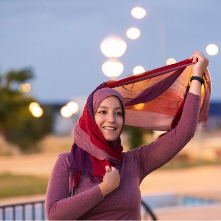 Eine junge Frau lächelt auf der Straße während sie eine Stoffstreifen ihres Kopftuchs in das Laternenlicht hält.