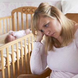 Mutter sitzt traurig vor einem Bettchen, in dem das Baby schläft: Nach der Geburt erleben viele Mütter einen Babyblues. Bleibt dieses Gefühl, haben sie eine Wochenbettdepression. Die Erkrankung kann jede Mutter treffen, bleibt aber oft unerkannt.