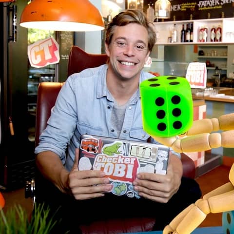 Checker Tobi sitzt hinter einer großen, hölzernen Spielfigur mit zwei großen Würfeln in blau und grün. | Bild: megaherz, Montage Tanja Begovic