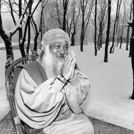 Bhagwan Shree Rajneesh hat die Hände wie zum Gebet zusammengelegt und lächelt. Er sitzt auf einem Stuhl in einer winterlichen Landschaft in Manali (Indien). Die Aufnahme ist vom 27.12.1985.