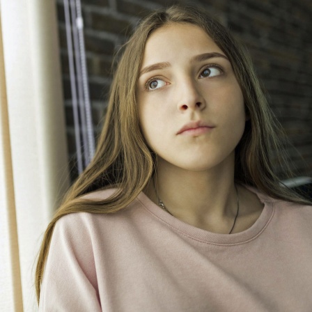 Ein Mädchen schaut traurig und sitzt zu Hause hinter einem Vorhang.