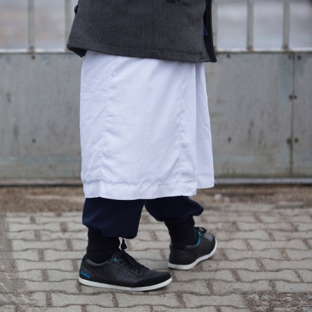 Ein Mann mit traditionellem weißen Gewand über Jogginghose und Turnschuhen steht vor einem Versammlungsort der Salafisten. Aufnahme von der Hüfte abwärts.