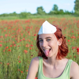 Mädchen mit Maske auf dem Kopf streckt in einem Mohnfeld fröhlich die Zunge heraus: