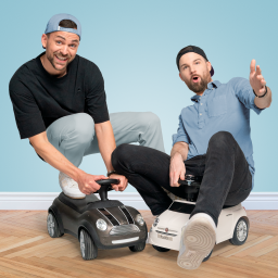 Die Bromance Daddys sitzen auf kleinen Kinderautos. Der Mann links trägt ein schwarzes T-Shirt, eine hellblaue Hose und eine umgedrehte blaue Basecap. Er lächelt und scheint Spaß zu haben. Der Mann rechts trägt ein hellblaues Hemd, schwarze Hosen und eine schwarze Basecap, die ebenfalls umgedreht ist. Er schaut überrascht oder begeistert. Beide sitzen auf kleinen, detailreichen Spielzeugautos, eines schwarz, das andere weiß. Der Hintergrund ist ein heller Blauton, und der Boden besteht aus Holzparkett.