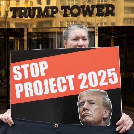 Eine grauhaarige Frau hält ein Schild mit dem Bild Trumps und der Aufschrift "Stop Project 2025". Hinter ihr der Eingang zum Gebäude mit der Aufschrift "Trump Tower".