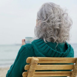 Grauhaarige Frau sitzt mit einer Tasse Tee auf einem Bambusstuhl am Strand 