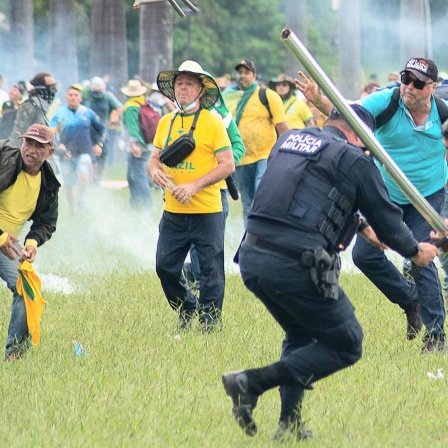 Ein Mann schlägt einen Polizisten mit einer Stange. Dahinter stürmen viele weitere Bolsonaro-Anhänger heran. 