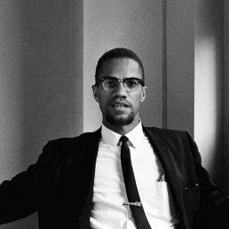 Malcolm X sitzt im Anzug und Krawatte im Foyer eines Hotels.