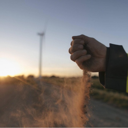 Ein Mann lässt Sand durch seine Hand rieseln, im Hintergrund ein Windrad auf einem Feld im Sonnenuntergang.
