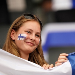 Zwei Mädchen halten lächelnd eine finnische Flagge hoch. Auf ihren Wangen sind ebenfalls finnische Flaggen gemalt.
