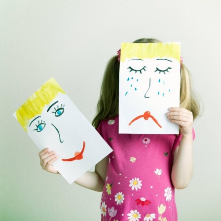 Ein Mädchen hält Zeichnungen mit einem lachendem und einem weinenden Gesicht in die Höhe.