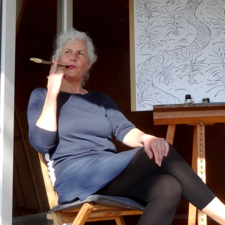 Miriam Wurster sitzt vor einer Leinwand mit einem Pinsel im Mund und schaut in die Ferne.
