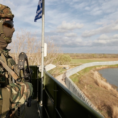 Ein vermummter Soldat bewacht mit Waffe von einer Erhöhung aus den Fluß Evros ind den umgebenden hohen Grenzzaun.