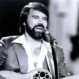 Country Sänger Glen Campbell steht mit seiner Gitarre an einem Mikro uns singt.
