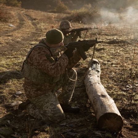 Zwei Soldaten knien hinter einem Baumstamm und schießen mit ihren Maschinengewehren.