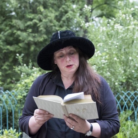 Annett Gröschner mit schwarzem Hut und schwarzem Oberteil liest vor grüner Kulisse aus einem Buch.
