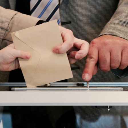 Ein Wahlbriefumschlag und die Hand eines Kindes, einer Frau und eines Mannes.