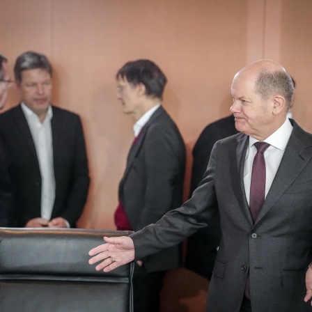 Bundeskanzler Olaf Scholz breitet die Arme aus, während sich im Hintergrund Wirtschaftsminister Robert Habeck und Gesundheitsminister Karl Lauterbach unterhalten
