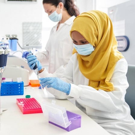 Zwei Wissenschaftlerinnen mit Maske, eine mit Hijab, arbeiten in einem Labor