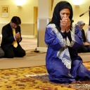Eine Frau mit schwarzem Kopftuch, in blauem Gewand kniet auf einem Gebetsteppich, hält die Augen geschlossen und die Hände vor das Gesicht. Hinter ihr knien in ähnlicher Haltung Männer und Frauen zum Gebet.