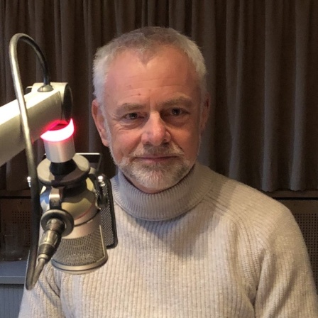 Thorsten Roos sitzt an einem Mikro in einem Radiostudio und blickt in die Kamera.