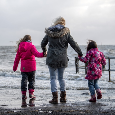 Eine Familie steht Hand in Hand bei Sturmflut an der Küste der Nordsee und schaut aufs Wasser: Bei Flut sind wir leichter und vom Erdmittelpunkt einen halben Meter weiter entfernt als bei Ebbe. Wie kann das sein?