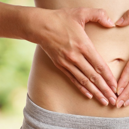 Eine Frau mit einem grauen Slip formt mit ihren Händen ein Herz und hält sie schützend vor ihren nackten Bauch.