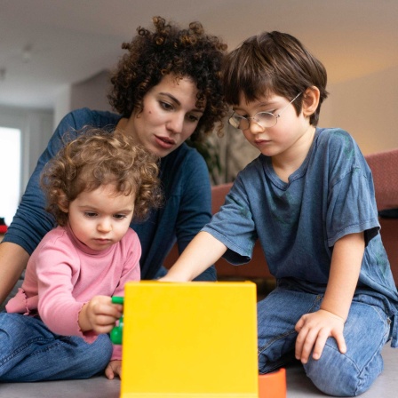 Symbolbild bezahlte Elternschaft: Eine Mutter spielt mit ihrern zwei Kindern; im Hintergrund der Vater.