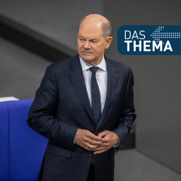 Olaf Scholz in einem dunklen Anzug, ein weißes Hemd und eine dunkle Krawatte. Im Hintergrund ist ein grauer Bereich mit einem blauen Stuhl sichtbar. Rechts oben im Bild befindet sich ein Textfeld mit der Aufschrift "DAS THEMA".