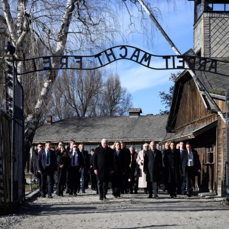 Eine Gruppe von dunkel gekleideten Menschen läuft durch das Tor in Auschwitz. Man kann den gespiegelten Schriftzug "Arbeit macht frei" über dem Tor lesen
