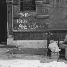 Ein Mann sitzt auf einem Fass vor einer Mauer auf der steht: The True America. 