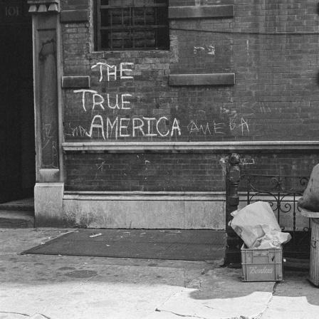 Ein Mann sitzt auf einem Fass vor einer Mauer auf der steht: The True America. 