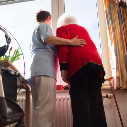 Eine Pflegerin (l) und eine Heimbewohnerin schauen zusammen aus einem Fenster im Wohnbereich des Pflegeheims. Im Vordergrund steht ein Rollator.