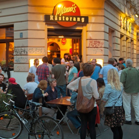 Symbolbild Lärm in der Nachbarschaft: Gäste stehen am Abend vor dem Restaurant und sitzen an Außentischen. 