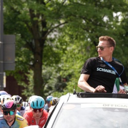 Der Ex-Radsportprofi André Greipel schaut aus dem Dachfenster eines fahrenden Autos auf Radprofis um ihn herum