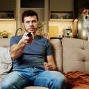 Ein Mann sitzt zu Hause auf einem Sofa und blickt Richtung Kamera; er hält eine Fernbedienung in der Hand. Auf dem Sofa steht ein Hund, der ebenfalls Richtung Kamera blickt.