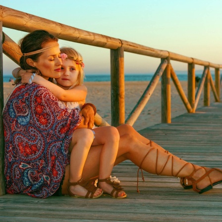Als Hippie gekleidete Mutter hält Tochter im Arm, am Strand