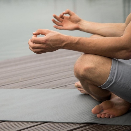 Ein Mann sitzt an einem Seeufer und meditiert auf einer Yogamatte 