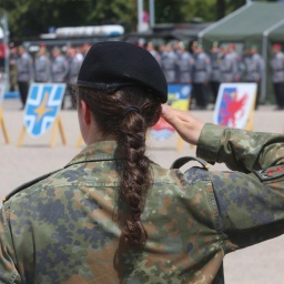 Eine Soldatin der Bundeswehr steht mit dem Rücken zum Bild. Sie hat einen schwarzen langen geflochtenen Zopf und hebt den rechten Arm nach oben und die Hand an die Stirn. Sie trägt eine Uniform. 